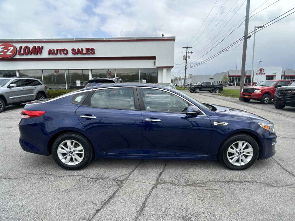 Blue sedan in a car lot with a dealership building in the background and power lines overhead.