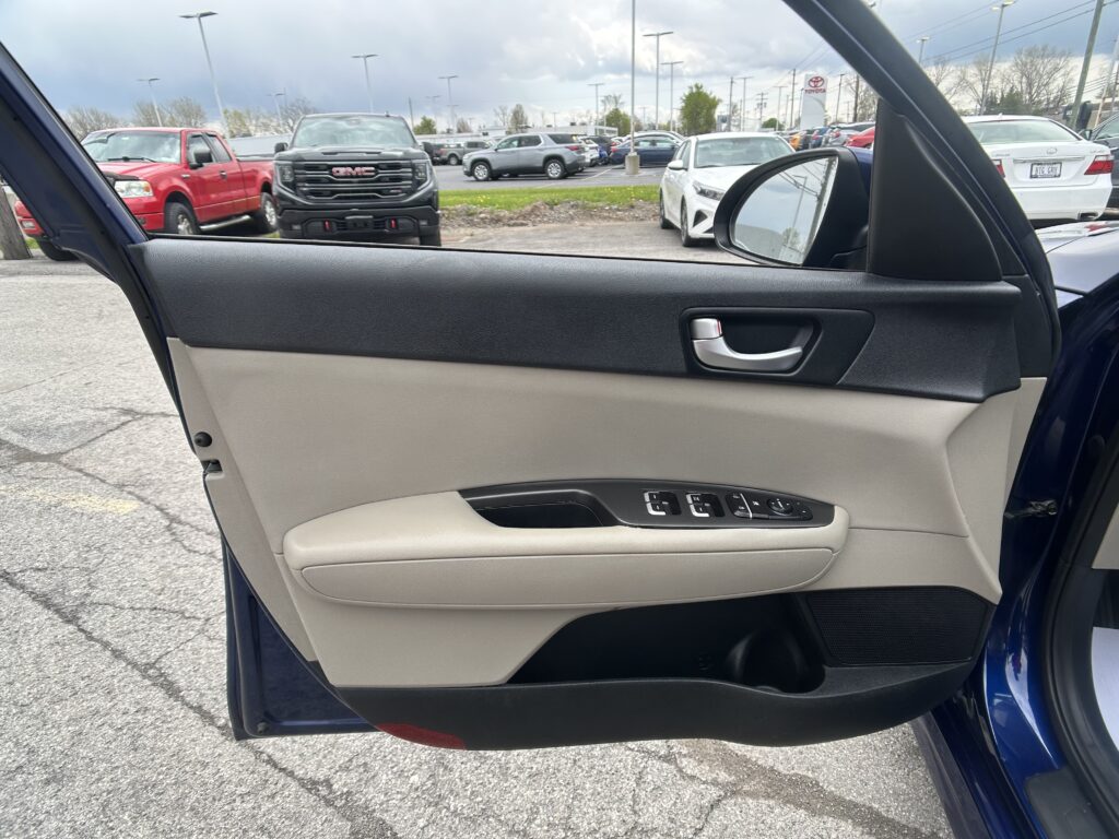 View of a car's open front passenger door from inside, showing the beige and black interior and parking lot outside with trucks and cars.