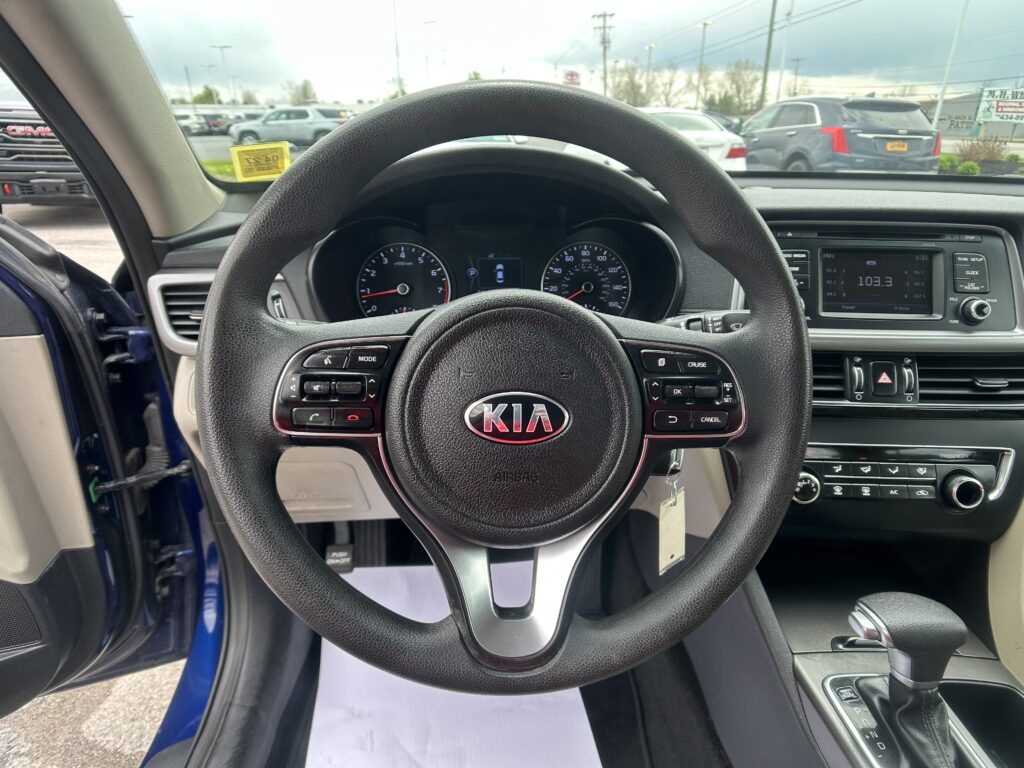 Driver's perspective of a Kia interior: steering wheel centered, dashboard gauges visible, radio on the right, parked in a lot outside the windshield.