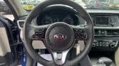Driver's perspective of a Kia interior: steering wheel centered, dashboard gauges visible, radio on the right, parked in a lot outside the windshield.