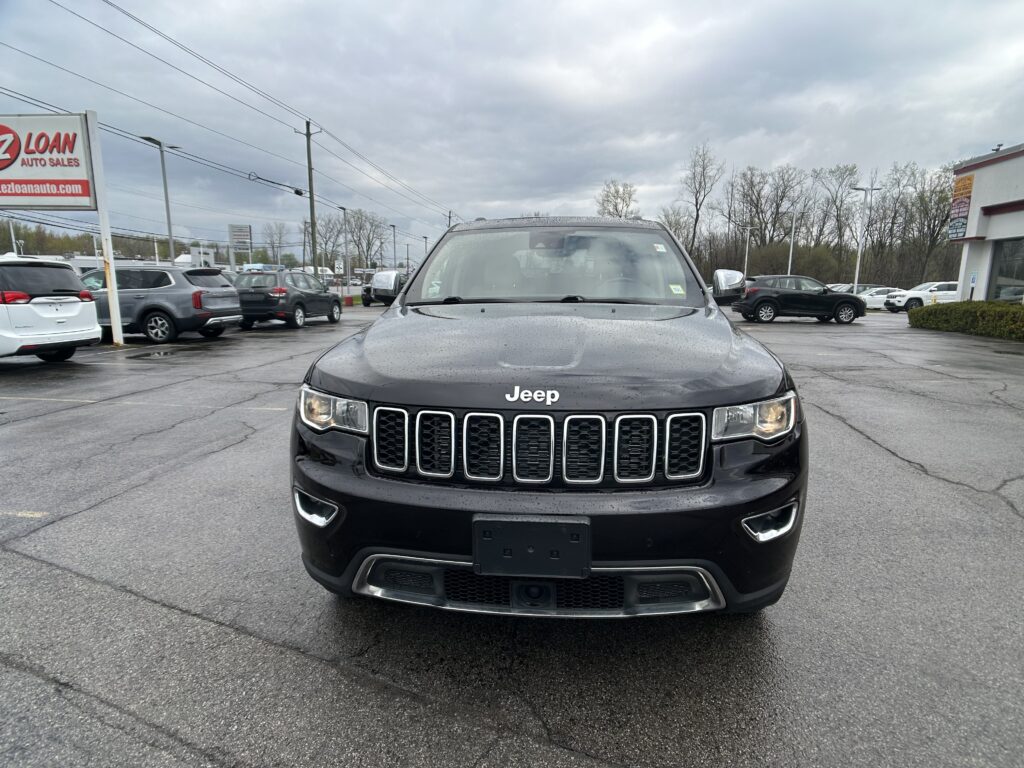 Front view of a black Jeep SUV parked in a dealership lot on a rainy day with other cars and a sign visible to the left.