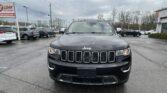 Front view of a black Jeep SUV parked in a dealership lot on a rainy day with other cars and a sign visible to the left.
