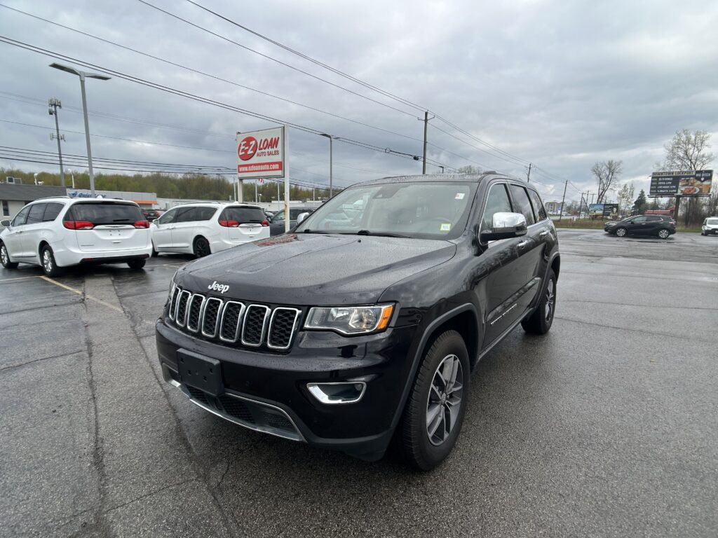 Dark gray Jeep SUV in a wet dealership lot, with white minivans in the background and an E-Z LOAN sign visible against a cloudy sky.