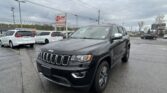 Dark gray Jeep SUV in a wet dealership lot, with white minivans in the background and an E-Z LOAN sign visible against a cloudy sky.
