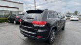 Rear view of a black Jeep Grand Cherokee in a parking lot, with other cars and a white building to the left under a cloudy sky.