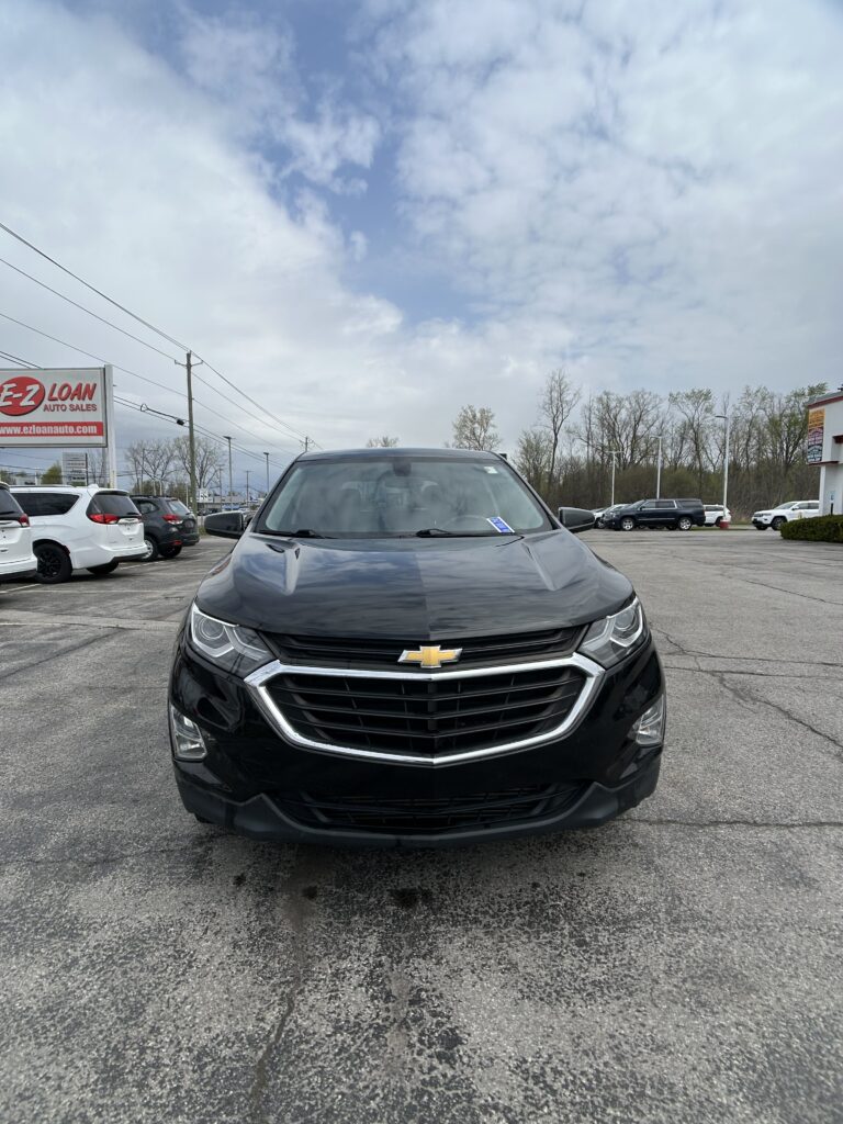 Front view of a black Chevrolet SUV parked in a dealership lot with other cars and a red-and-white EZ Loan Auto Sales sign in the background.