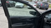 Open car door in the foreground with a busy dealership parking lot in the background on a cloudy day, including a white car nearby and a Toyota sign farther away.