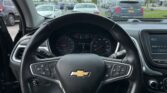 View from the driver's seat of a Chevrolet, showing the steering wheel and dashboard with a row of cars outside on a cloudy day in a parking lot.