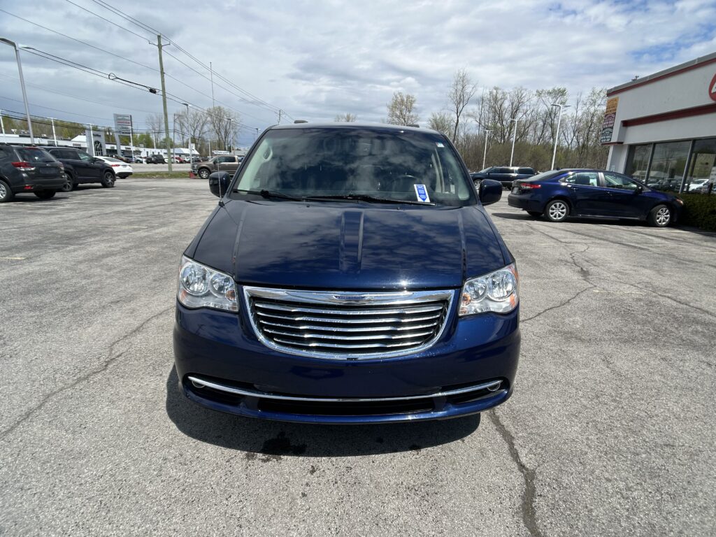 Front view of a dark blue SUV in a parking lot near a car dealership, with other vehicles and a building in the background under a cloudy sky.