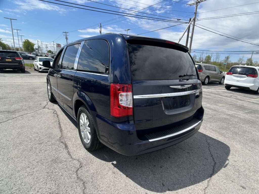 Rear three-quarter view of a dark blue Town & Country minivan parked in a busy car lot with other vehicles and power lines overhead.