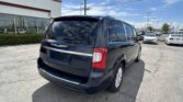 Rear view of a dark blue Chrysler minivan in a parking lot next to a white building with glass storefronts, blue sky above.