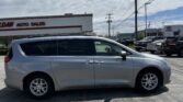 Silver minivan parked in a car dealership lot with other vehicles and a dealership building in the background under a cloudy sky.