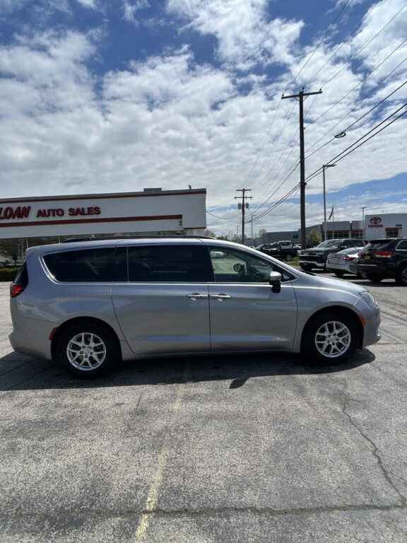 Silver minivan parked in a car dealership lot with other vehicles and a dealership building in the background under a cloudy sky.