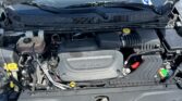Open car hood showing engine bay with coolant reservoir, air intake, battery, and windshield wipers visible; front grille in foreground.