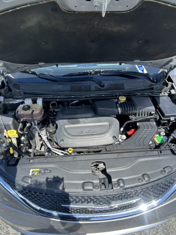 Open car hood showing engine bay with coolant reservoir, air intake, battery, and windshield wipers visible; front grille in foreground.