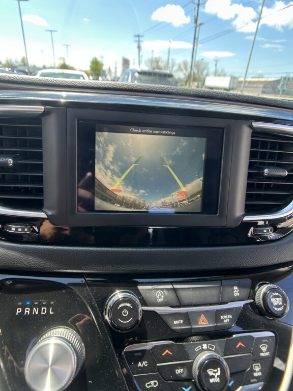 In-dash backup camera view with yellow parking guide lines on a sunny parking lot backdrop, center screen on a car dashboard