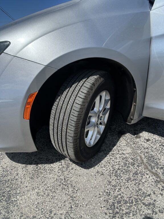Front left wheel of a silver car, showing the tire, alloy rim, and wheel well with an orange side marker light beside the fender on asphalt.