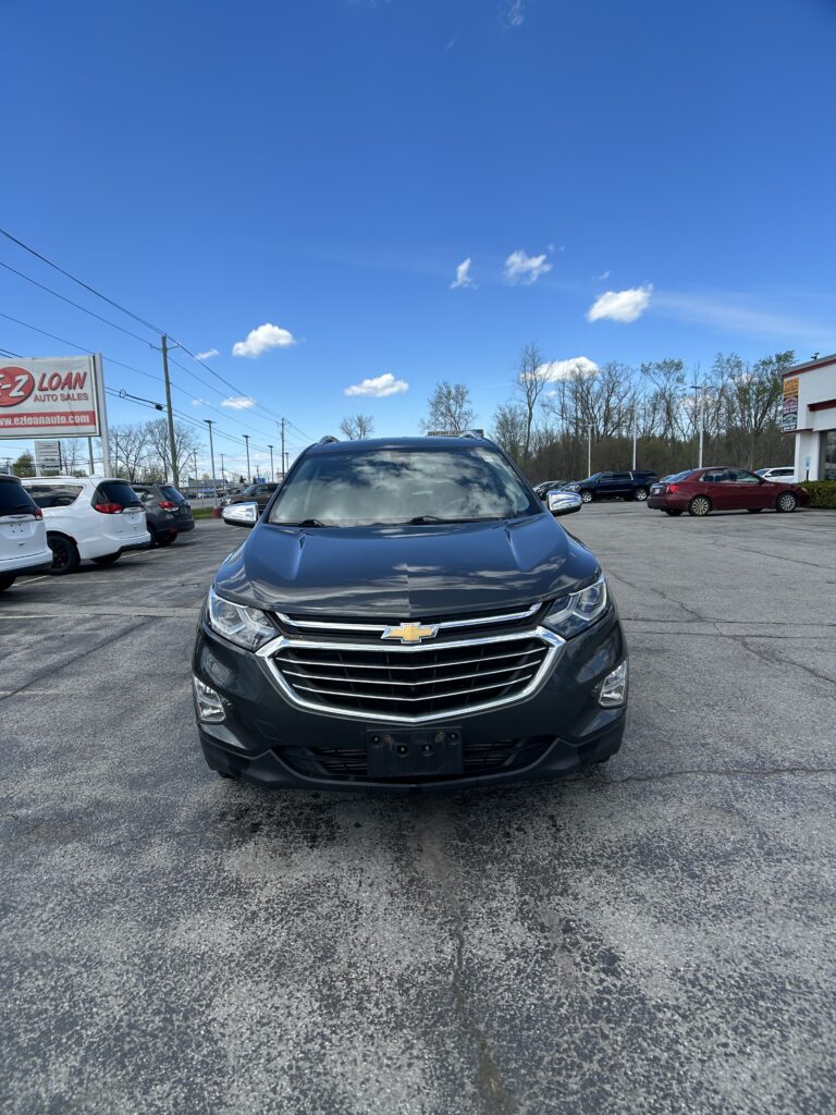 Front view of a dark gray Chevrolet SUV parked in a car dealership lot under a clear blue sky, with other vehicles nearby and a dealership sign visible to the left.