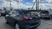 Rear three-quarter view of a dark gray Chevrolet Equinox SUV parked in a lot with other cars, power lines overhead and a blue sky with clouds in the background.