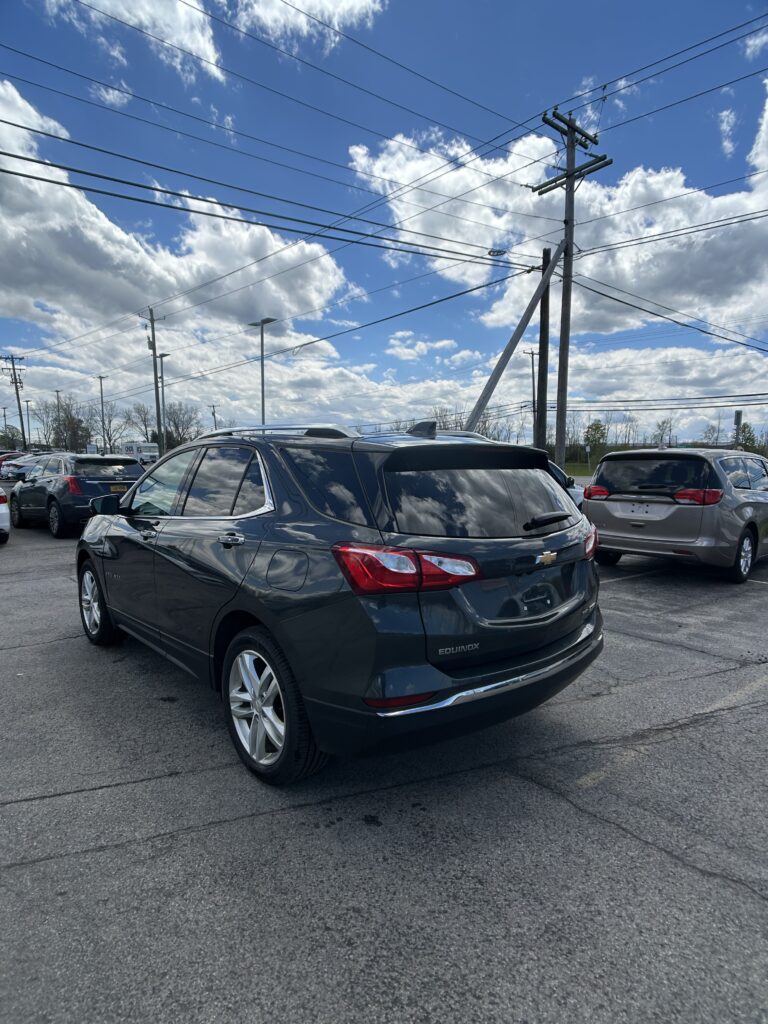 Rear three-quarter view of a dark gray Chevrolet Equinox SUV parked in a lot with other cars, power lines overhead and a blue sky with clouds in the background.