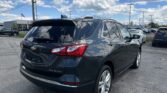 Rear three-quarter view of a dark gray Chevrolet SUV in a car dealership lot on a sunny day with power lines overhead and a blue sky.