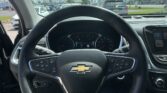 Inside a car cockpit viewed from the driver’s seat: steering wheel with Chevy emblem and instrument cluster, with a parking lot and blue sky outside the windshield.