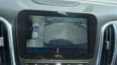 View from inside a car showing the center touchscreen with a rearview camera feed of a parking lot and nearby white and dark vehicles behind, plus guiding colored lines on the screen.