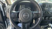Driver's-seat view of a Jeep dashboard: steering wheel, gauges, center console, and an open driver’s door; parking lot outside.