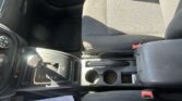 Close-up view of a car's front interior showing the center console, gear shifter, cupholders, and a two-tone fabric seat with sunlight creating shadows; a sheet of paper rests on the floor.