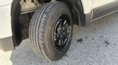 Close-up of a light-colored SUV's front wheel and tire in the wheel well, showing a black alloy rim and exposed suspension.