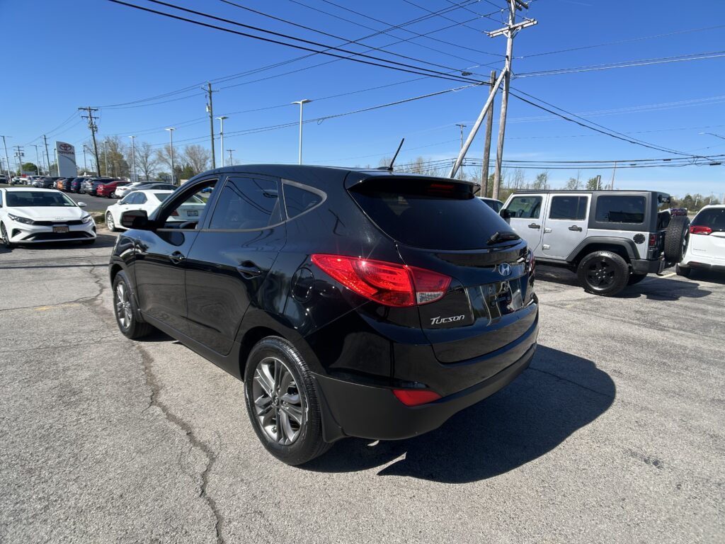Rear three-quarter view of a black Hyundai Tucson in a car dealership lot, with other cars and a Toyota sign in the distance under a clear blue sky.