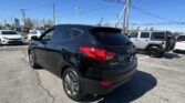 Rear three-quarter view of a black Hyundai Tucson in a car dealership lot, with other cars and a Toyota sign in the distance under a clear blue sky.