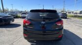 Rear view of a black Hyundai Tucson in a crowded parking lot under a clear blue sky, with other cars nearby.