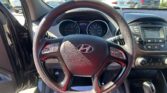 Front-seat view of a car’s steering wheel and dashboard, with a parking lot seen through the windshield behind. Short focus on speedometer and center console buttons in the foreground.