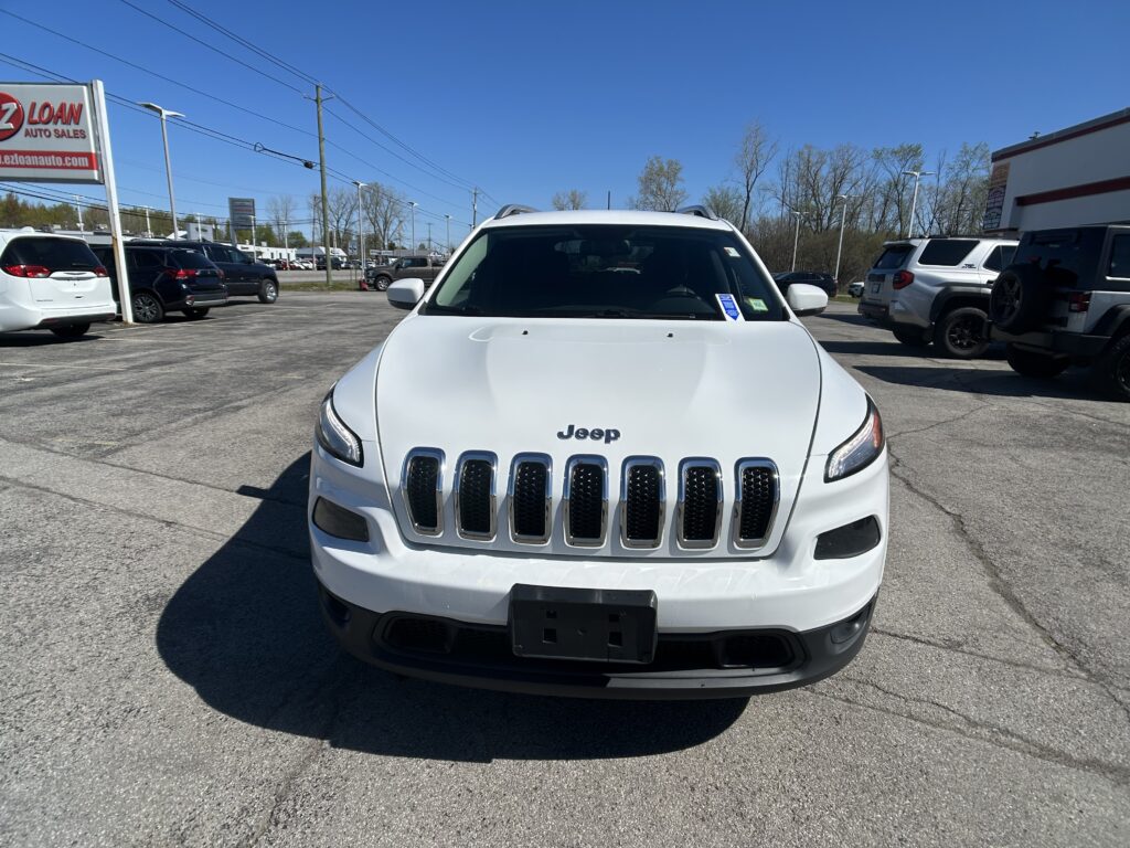Front view of a white Jeep SUV parked in a dealership lot with other cars and a bright blue sky above.",