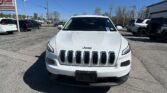 Front view of a white Jeep SUV parked in a dealership lot with other cars and a bright blue sky above.",