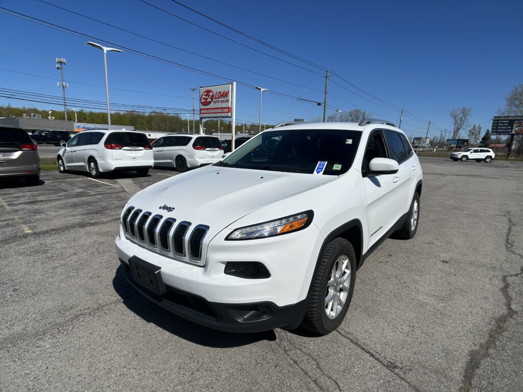 White Jeep SUV in the foreground at a car dealership lot, with several white vans and an EZ Loan Auto Sales sign under a clear blue sky.