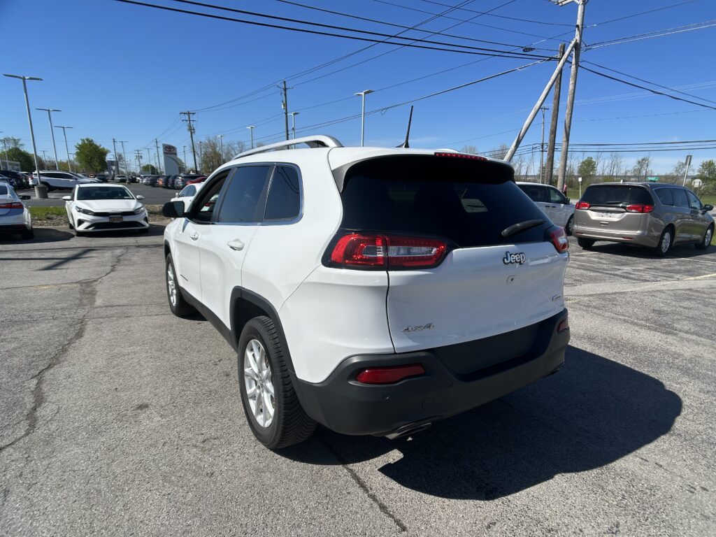 White Jeep SUV in a dealership parking lot with other cars and a clear blue sky above.