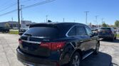 Rear three-quarter view of a black Acura MDX SUV in a dealership parking lot under a clear blue sky, with power lines overhead.