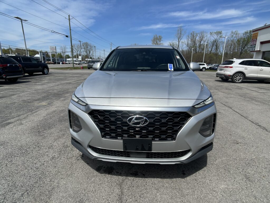 Close-up, front view of a silver Hyundai SUV parked in a busy car lot on a sunny day, dealership buildings in the background