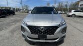 Close-up, front view of a silver Hyundai SUV parked in a busy car lot on a sunny day, dealership buildings in the background