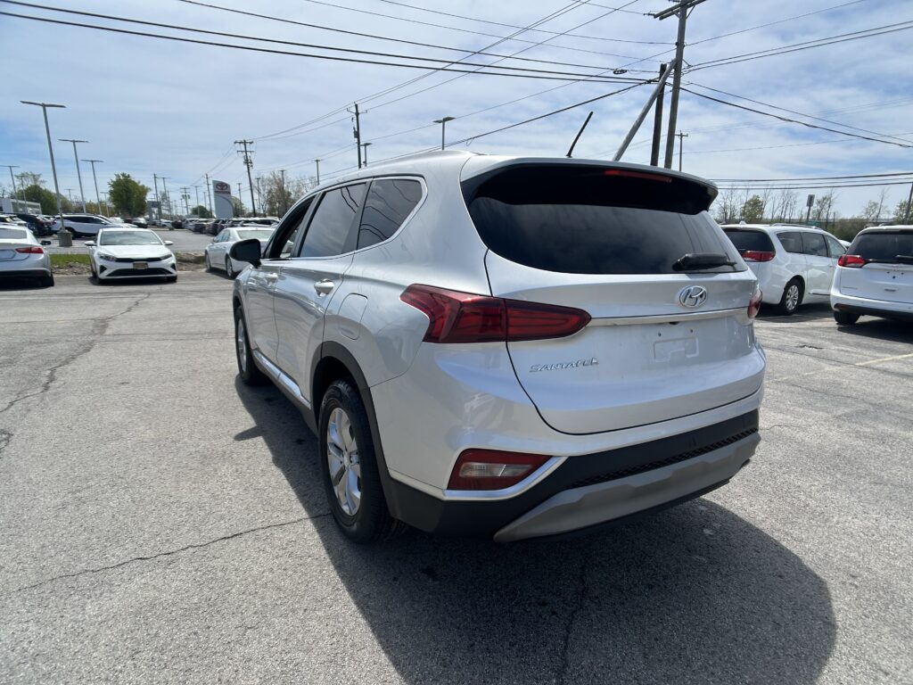 Rear three-quarter view of a white Hyundai Santa Fe SUV in a car dealership lot, with other vehicles and power lines in the background.