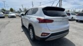 Rear three-quarter view of a white Hyundai Santa Fe SUV in a car dealership lot, with other vehicles and power lines in the background.