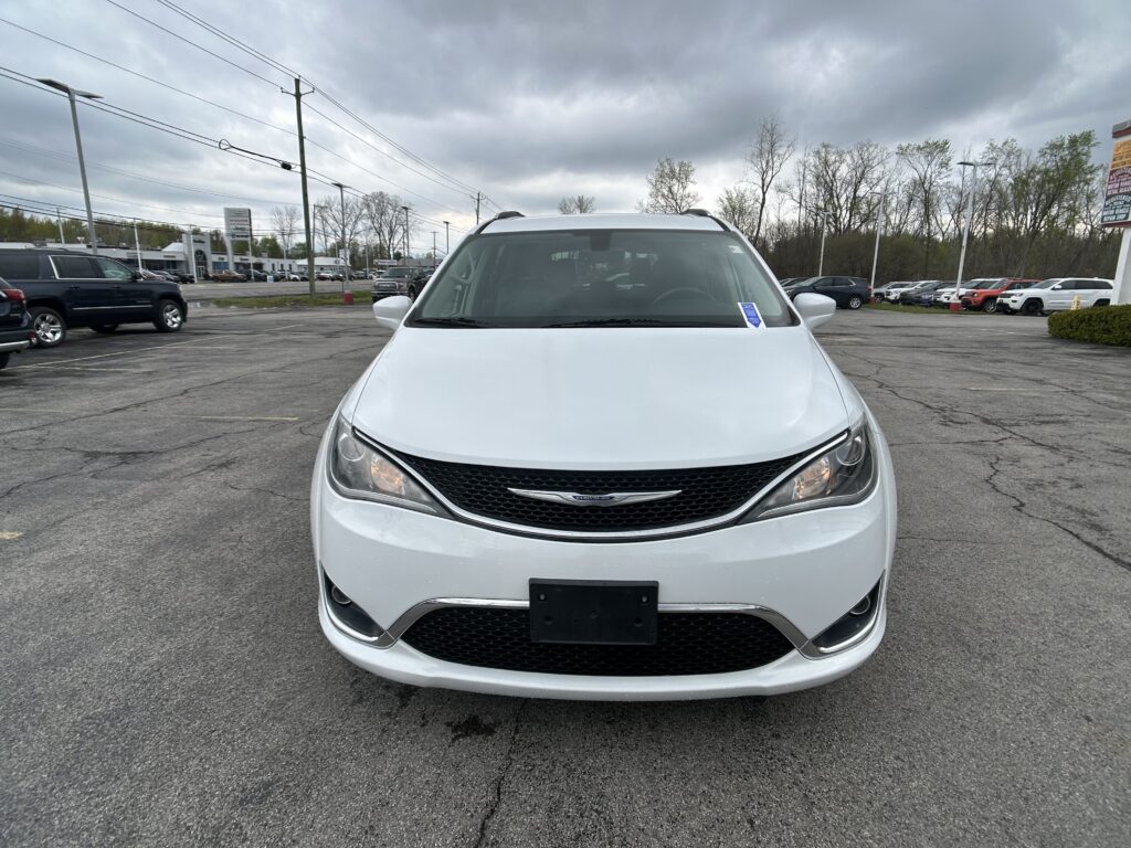 Front view of a white Chrysler minivan parked in a crowded lot under a cloudy sky, with other cars in the background.