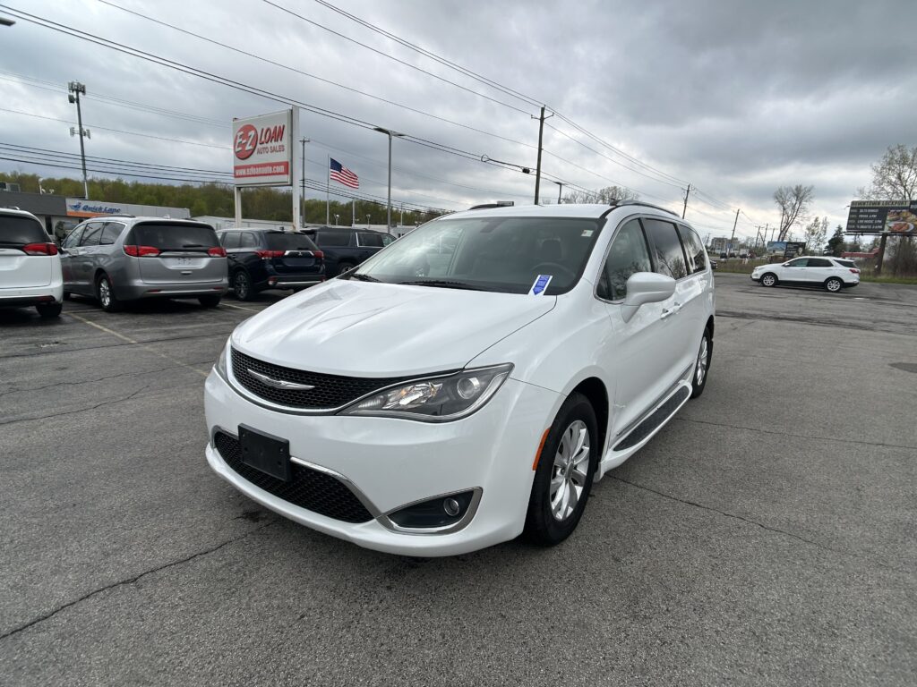 White Chrysler minivan in the foreground at a used-car lot with an EZ Loan Auto Sales sign and an American flag under a cloudy sky.