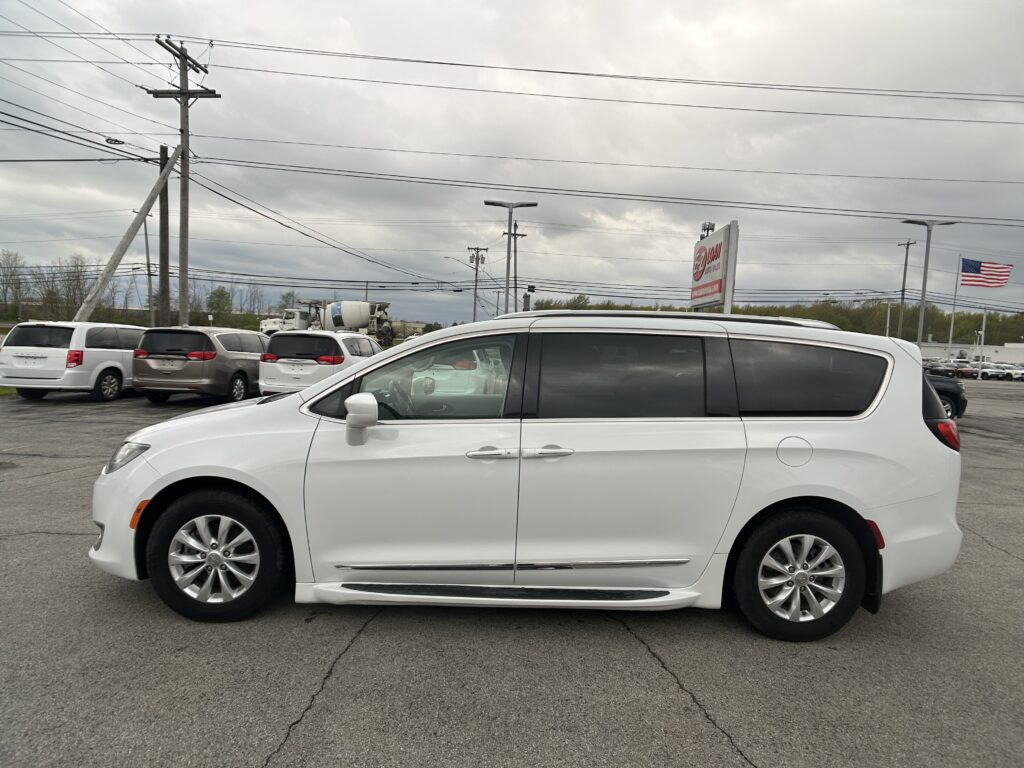 White minivan in a parking lot with other vehicles, power lines overhead, and an overcast sky; a flag and dealership sign visible in the distance.