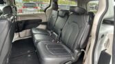 Interior of a SUV or minivan showing black leather seats with red stitching in the second row, beige door panels, and seat belts visible; outside windows show other parked vehicles and a dealership lot.