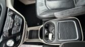 Close-up of a car's center console with climate controls, gear selector, cup holders, and a perforated black leather seat nearby.