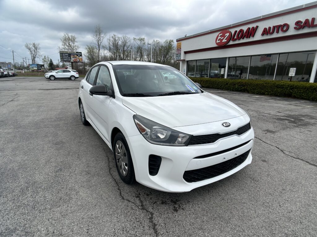 White Kia sedan in the foreground at an auto dealer lot, with EZ Loan Auto Sales storefront and large glass windows in the background under a cloudy sky.