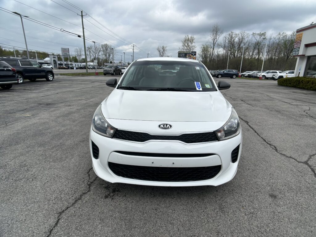 Front view of a white Kia car parked in a dealership lot on a cloudy day, with other vehicles in the background and storefronts visible far away.